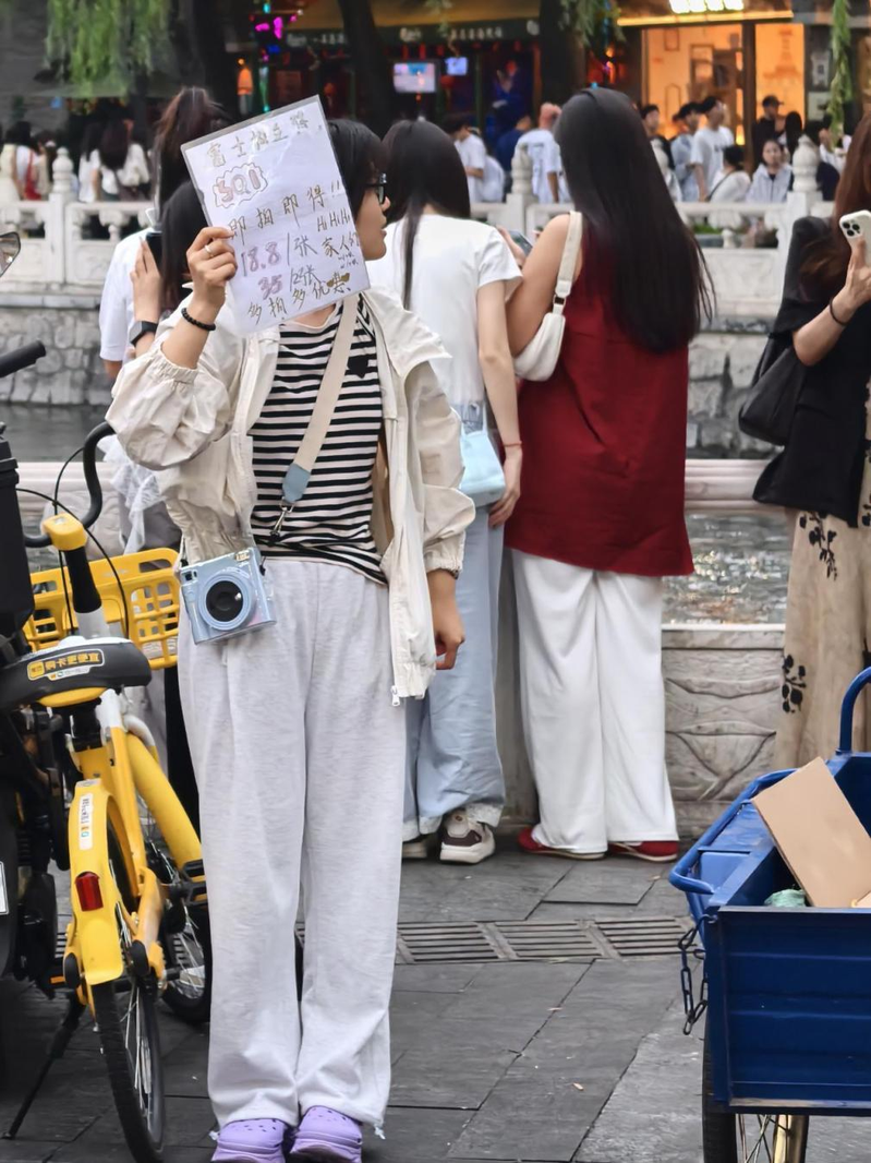 一名女子手里举着手写的广告牌和客片,胸前挂着微信支付收款码,胳膊上挂着拍立得相机,正冲着过往人群招揽生意。(取材自北京日报)