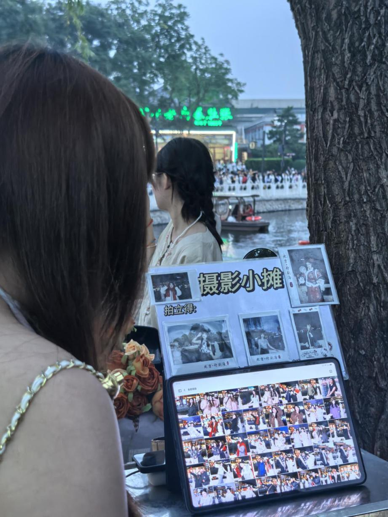 一名女子手里举着手写的广告牌和客片，胸前挂着微信支付收款码，胳膊上挂着拍立得相机，正冲着过往人群招揽生意。（取材自北京日报）