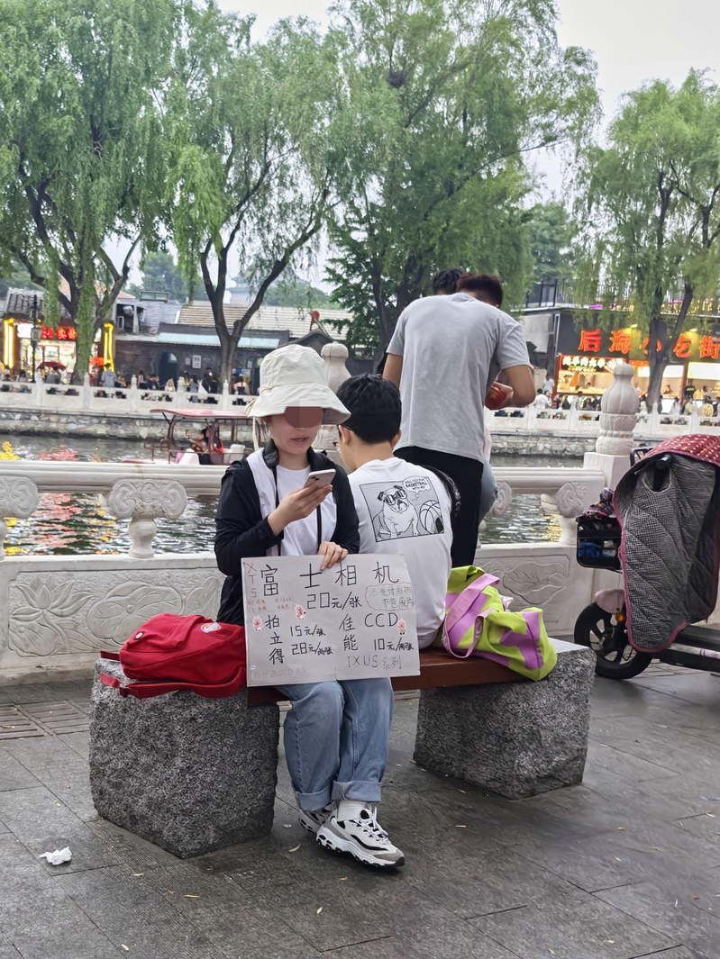 一名女子手里举着手写的广告牌和客片，胸前挂着微信支付收款码，胳膊上挂着拍立得相机，正冲着过往人群招揽生意。（取材自北京日报）