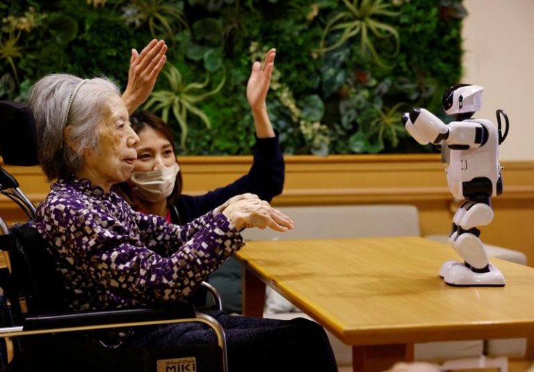 在日本东京一间由善光会经营的养老设施，居民行方幸子（左）和她的照服员跟随一台名为PALRO的小型人形机器人做动作。（路透）