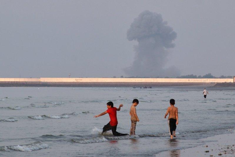 三名男孩13日在伊朗阿巴斯港外海的荷莫兹海峡浅水区玩耍,远处可见爆炸升起的烟柱。...