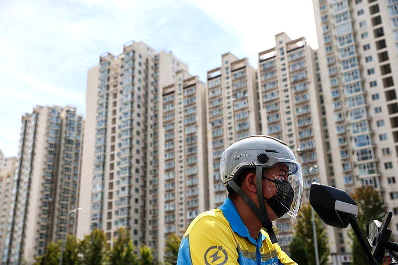 一名外送员骑着电动车经过高楼大厦。2026年7月,中国70个大中城市新建商品住宅...
